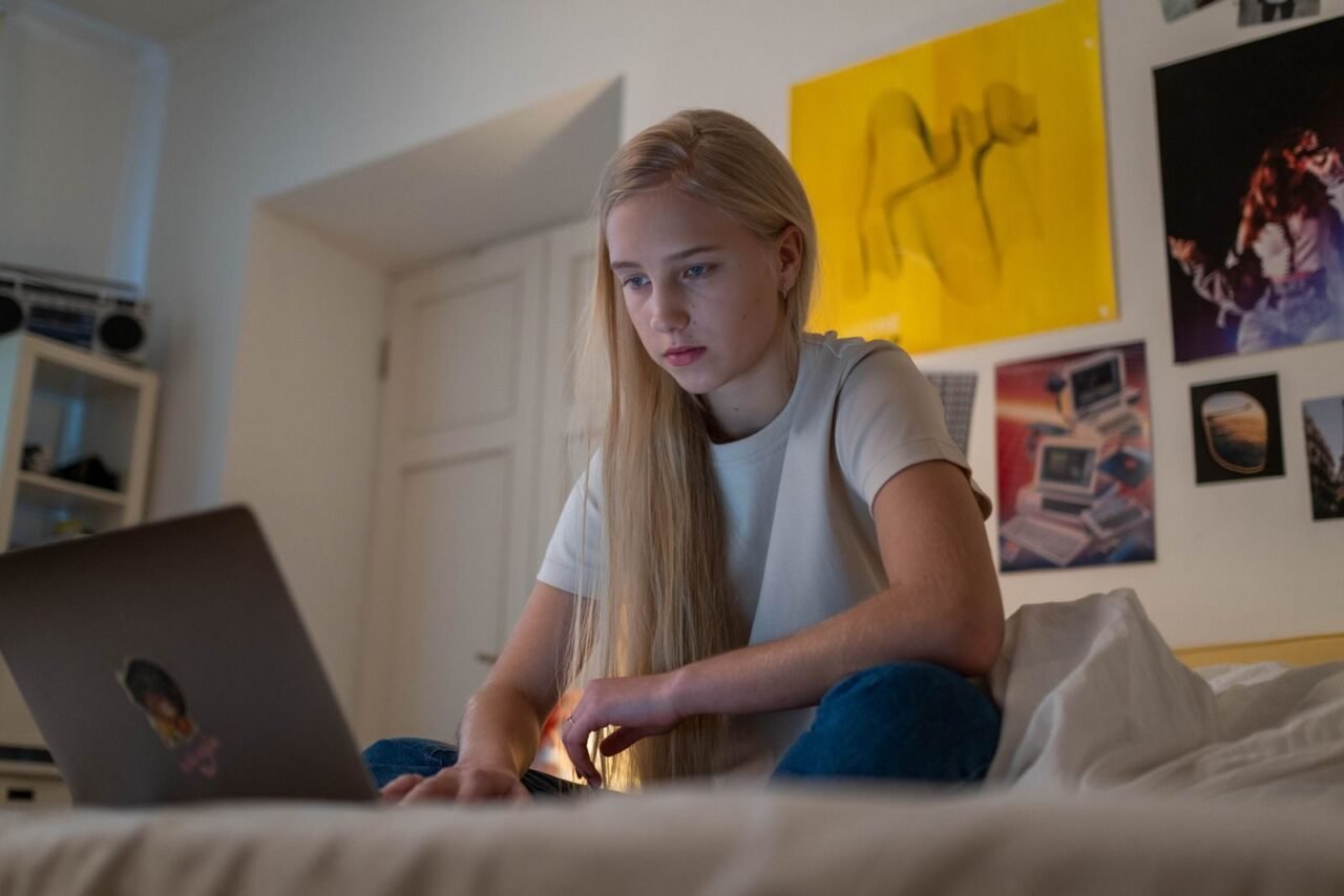 A focused teenage girl with long blonde hair using a laptop in her bedroom for study or leisure activities.