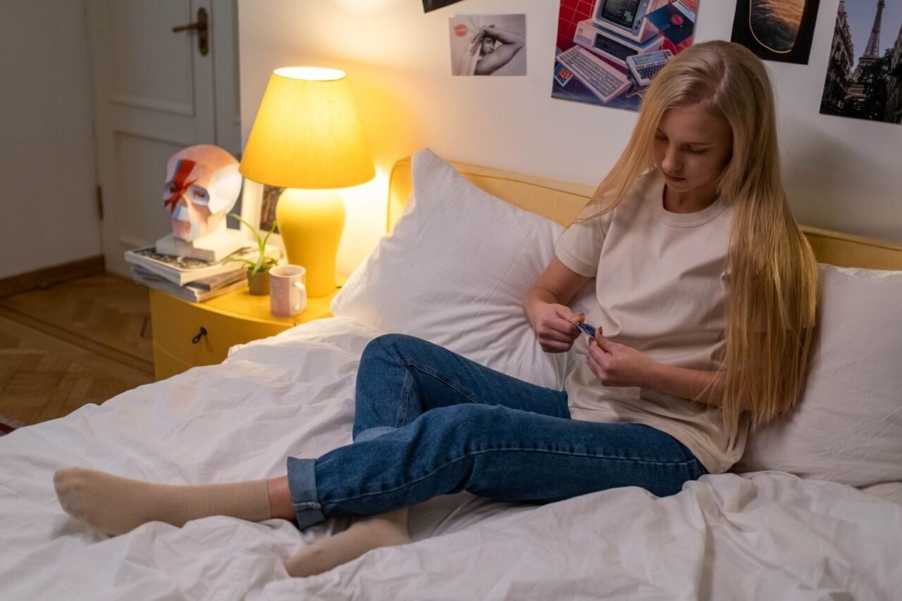 Teen girl in a bedroom setting, holding a contraceptive item, on a bed with soft lighting.