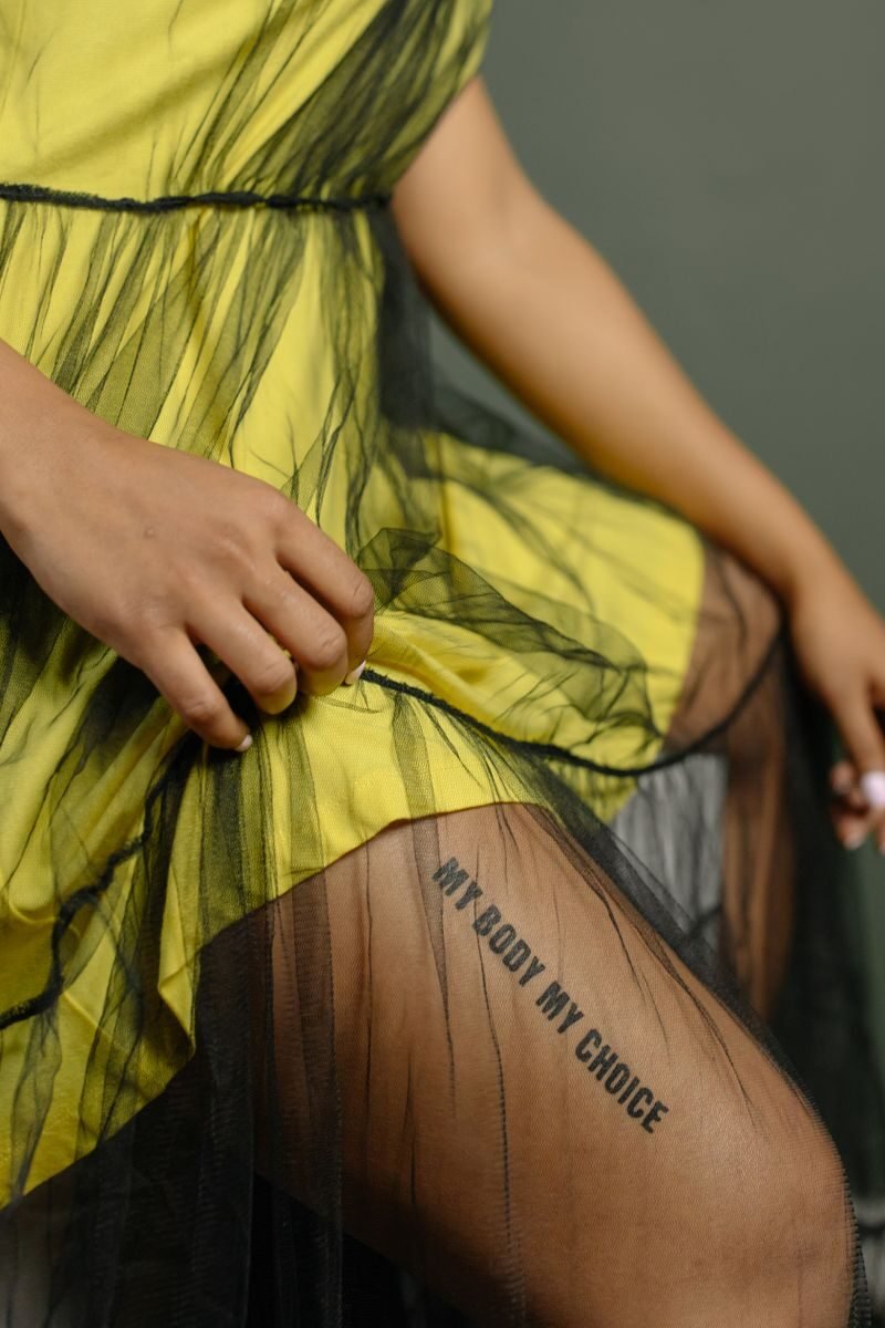 Close-up of female leg with 'My Body My Choice' tattoo, wearing yellow dress.