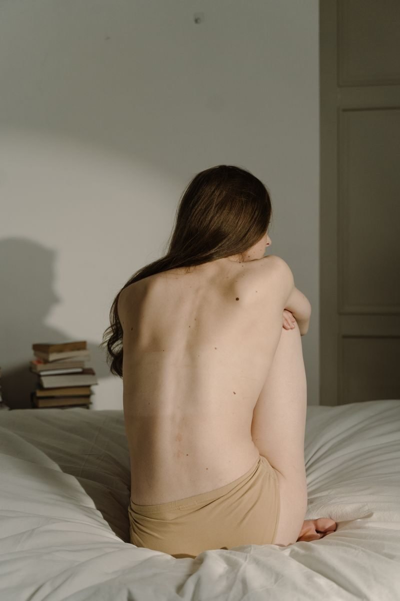 A woman is sitting on a bed, showing her back, in a calming indoor setting.