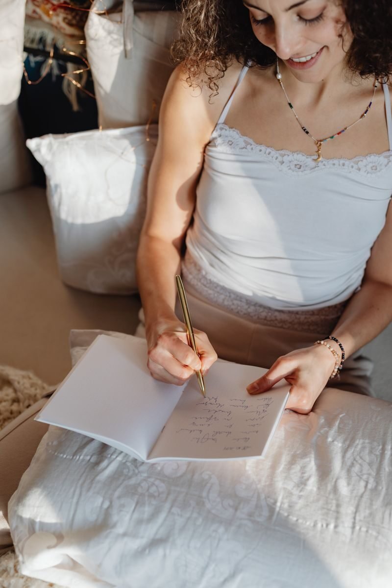 A brunette woman writes and smiles while sitting comfortably indoors with a notebook.