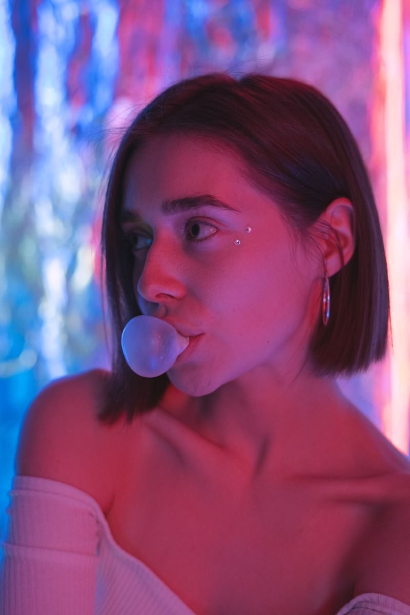 A young woman blows a bubble with gum, illuminated by vibrant colored lights. Side profile view.