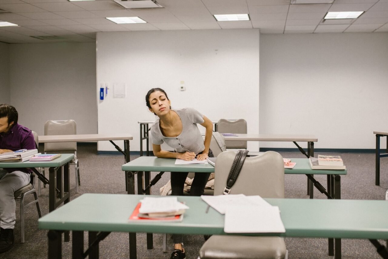 A college student appears to be cheating during an exam in a classroom setting, bending over to glance at another student's paper.