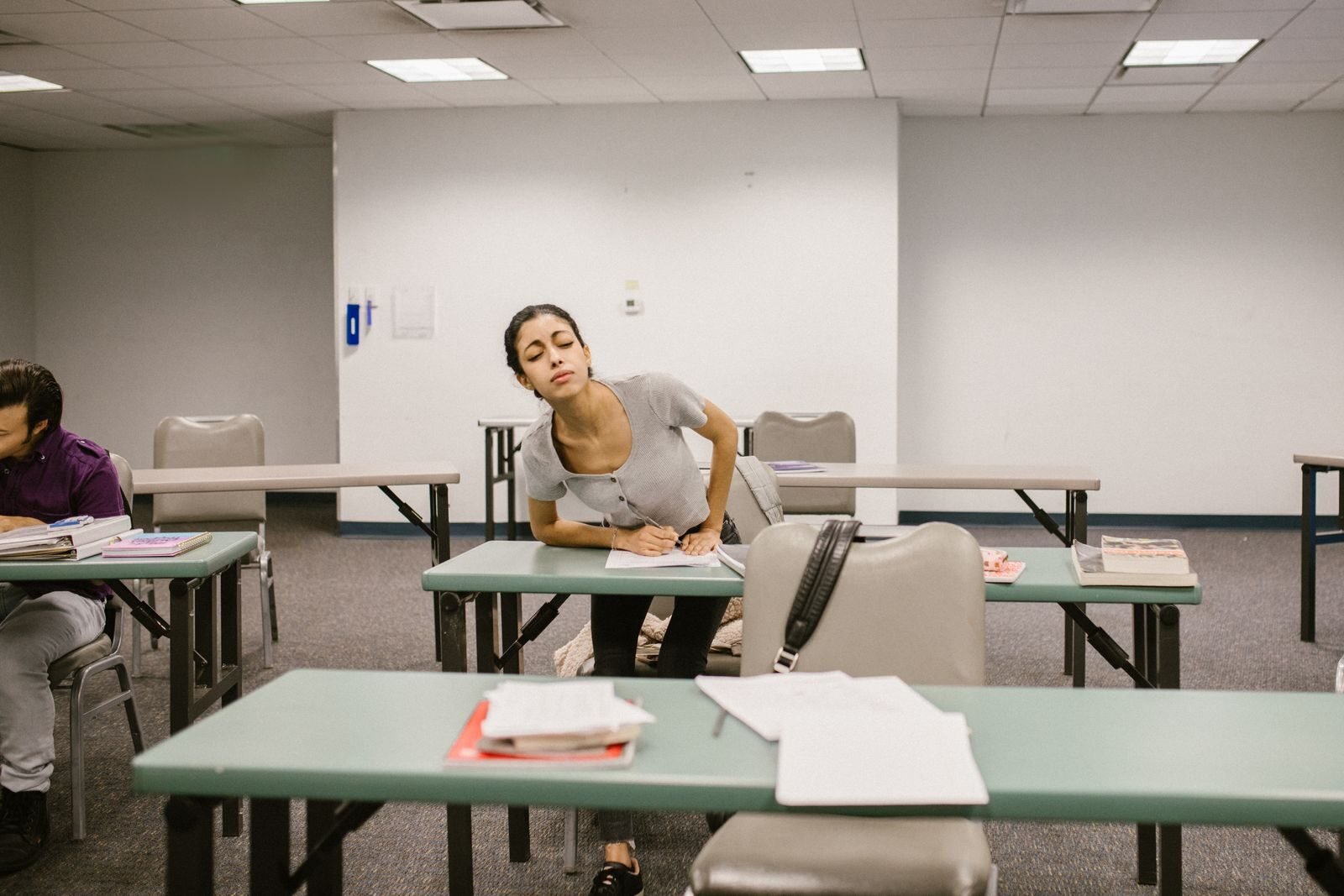 A college student appears to be cheating during an exam in a classroom setting, bending over to glance at another student's paper.
