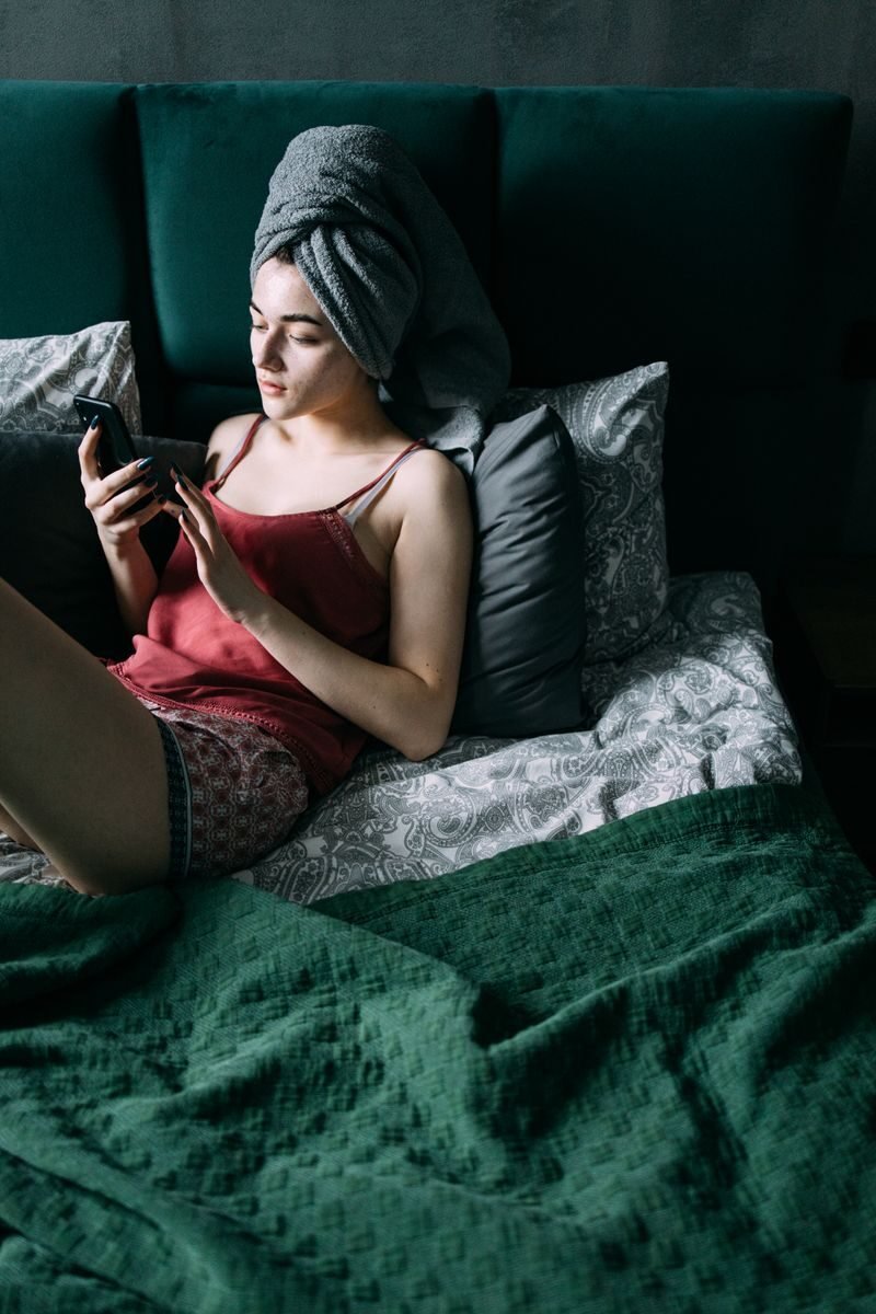 Woman in bed with hair wrapped in towel, using smartphone, under green blanket.
