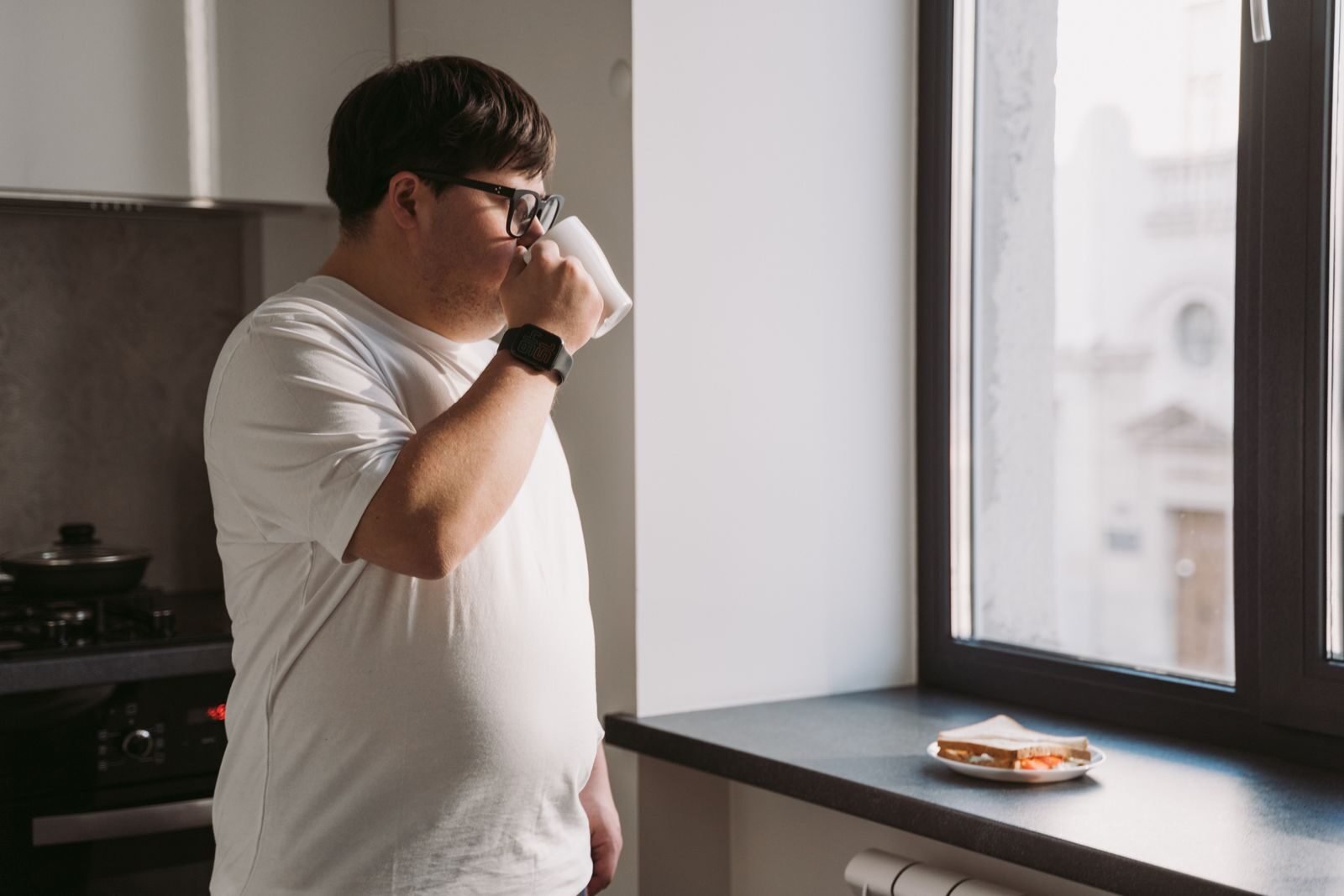 Man with Down Syndrome drinking coffee by the window in a kitchen setting.