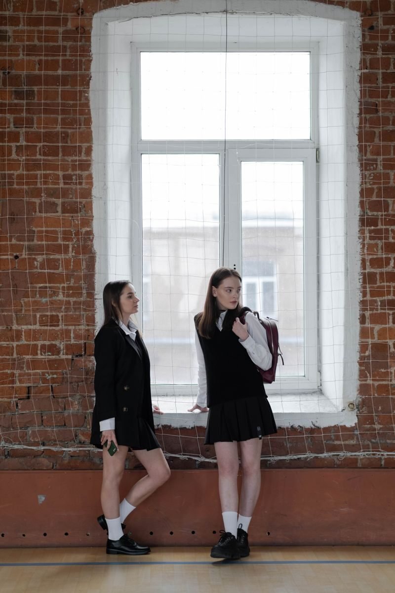 Two teenage girls in school uniforms standing by a brick wall and window indoors.