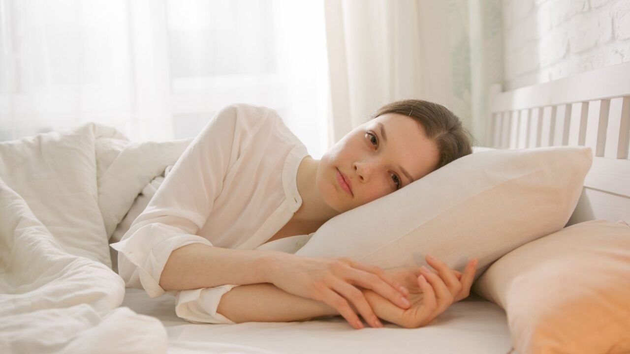 A woman in a white shirt lies on her side in bed, embracing a pillow, framed by soft natural lighting.