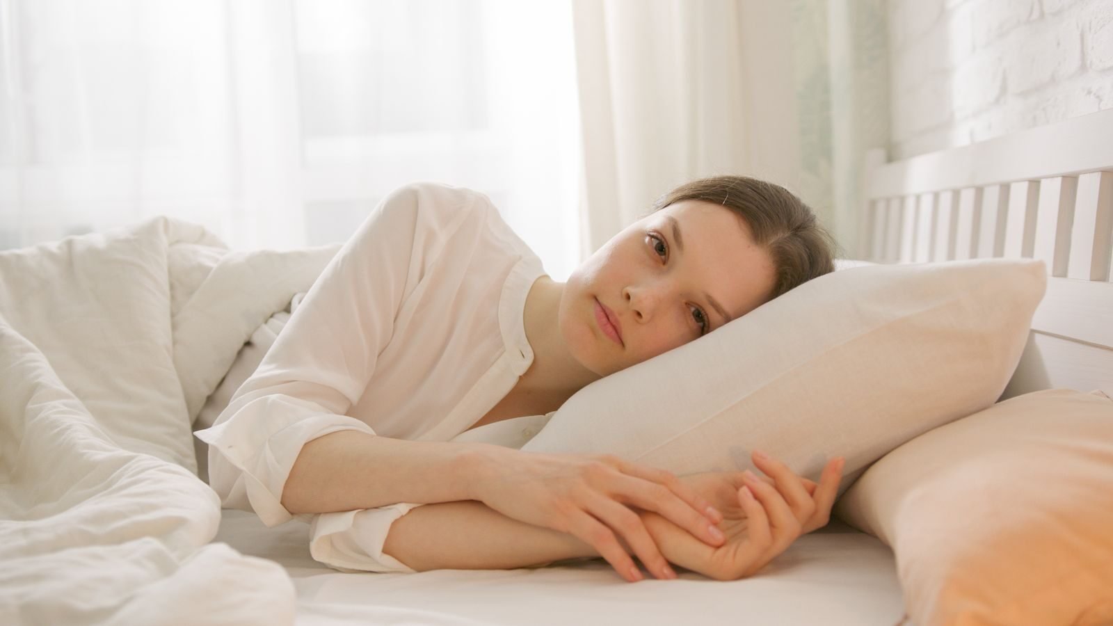 A woman in a white shirt lies on her side in bed, embracing a pillow, framed by soft natural lighting.