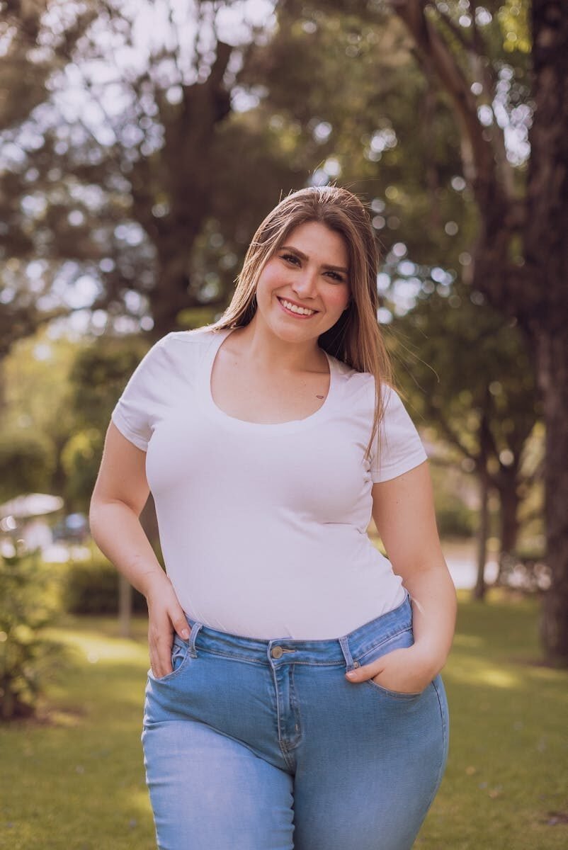 A cheerful woman standing in a park, wearing a white shirt and jeans.