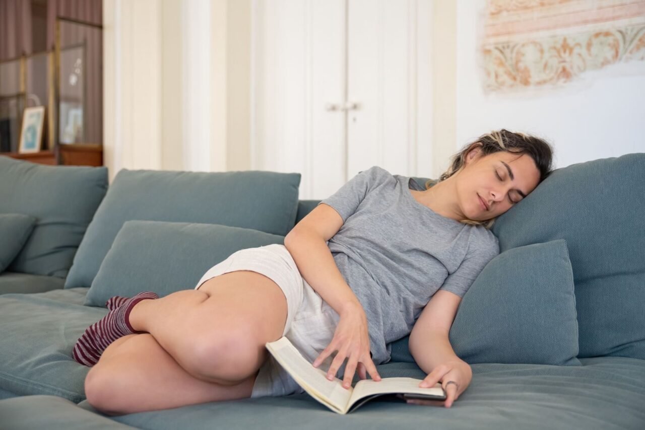 Woman peacefully napping on a cozy gray sofa with an open book in a serene interior setting.