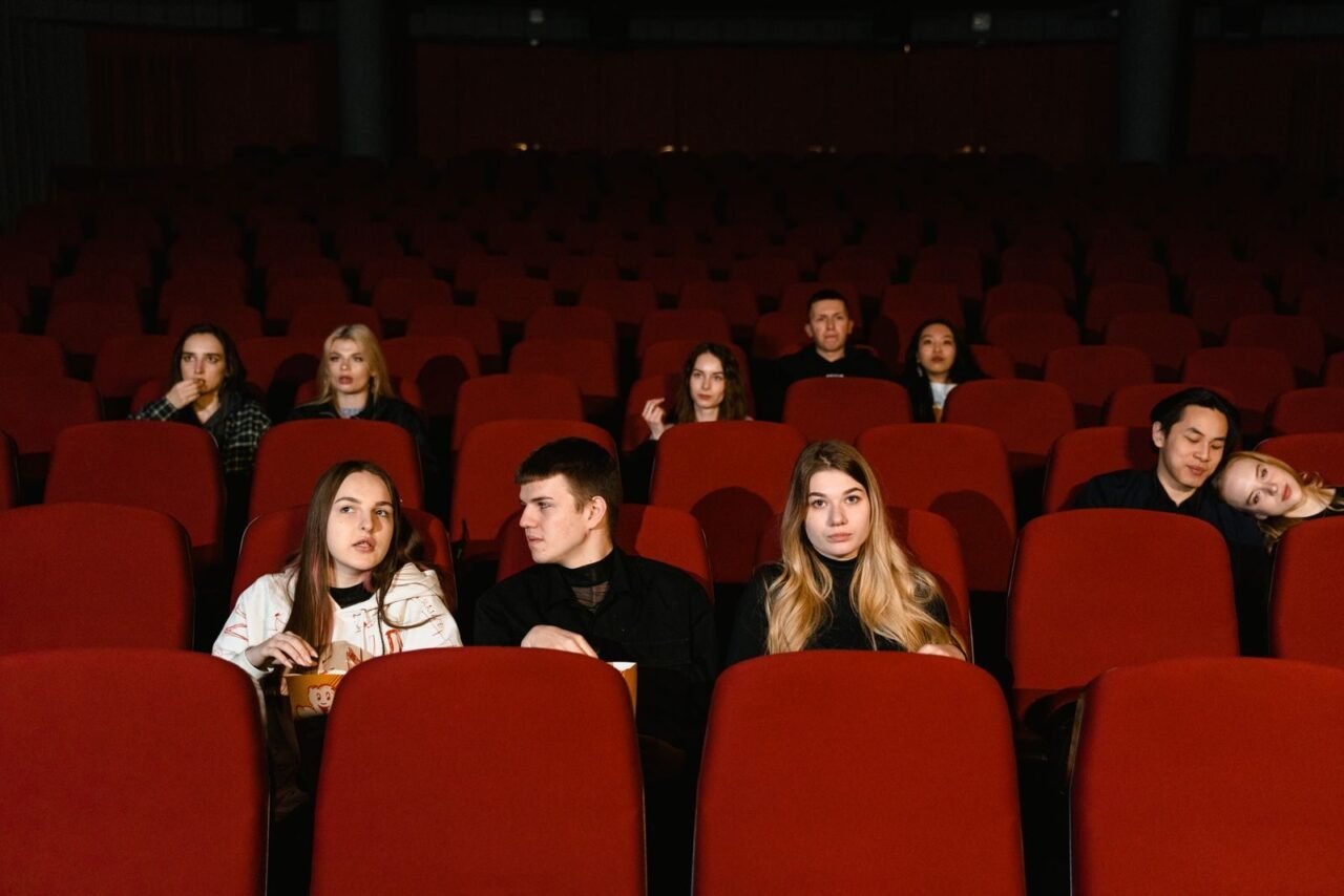 A diverse group of adults seated in a cinema, watching a movie with red seats and popcorn.