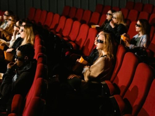 People wearing 3D glasses and eating popcorn in a cinema with red chairs.