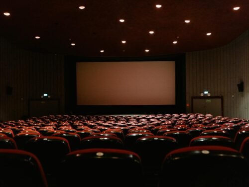 A dimly lit empty cinema theater with rows of red chairs facing a projector screen.