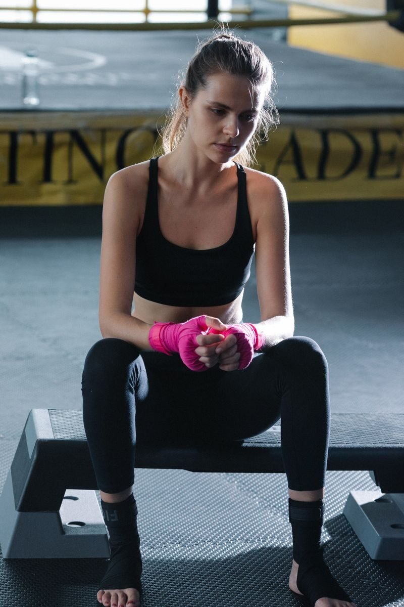 A female boxer with pink gloves sits on a bench, taking a break in a gym setting.