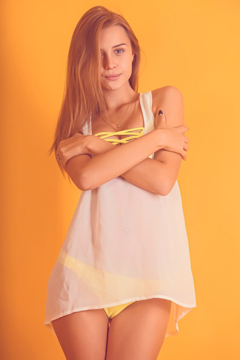 Confident woman in swimsuit posing with arms crossed against a bright orange backdrop.