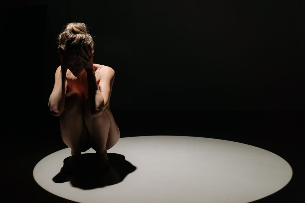 A woman crouching under a spotlight with hands on head, depicting stress.