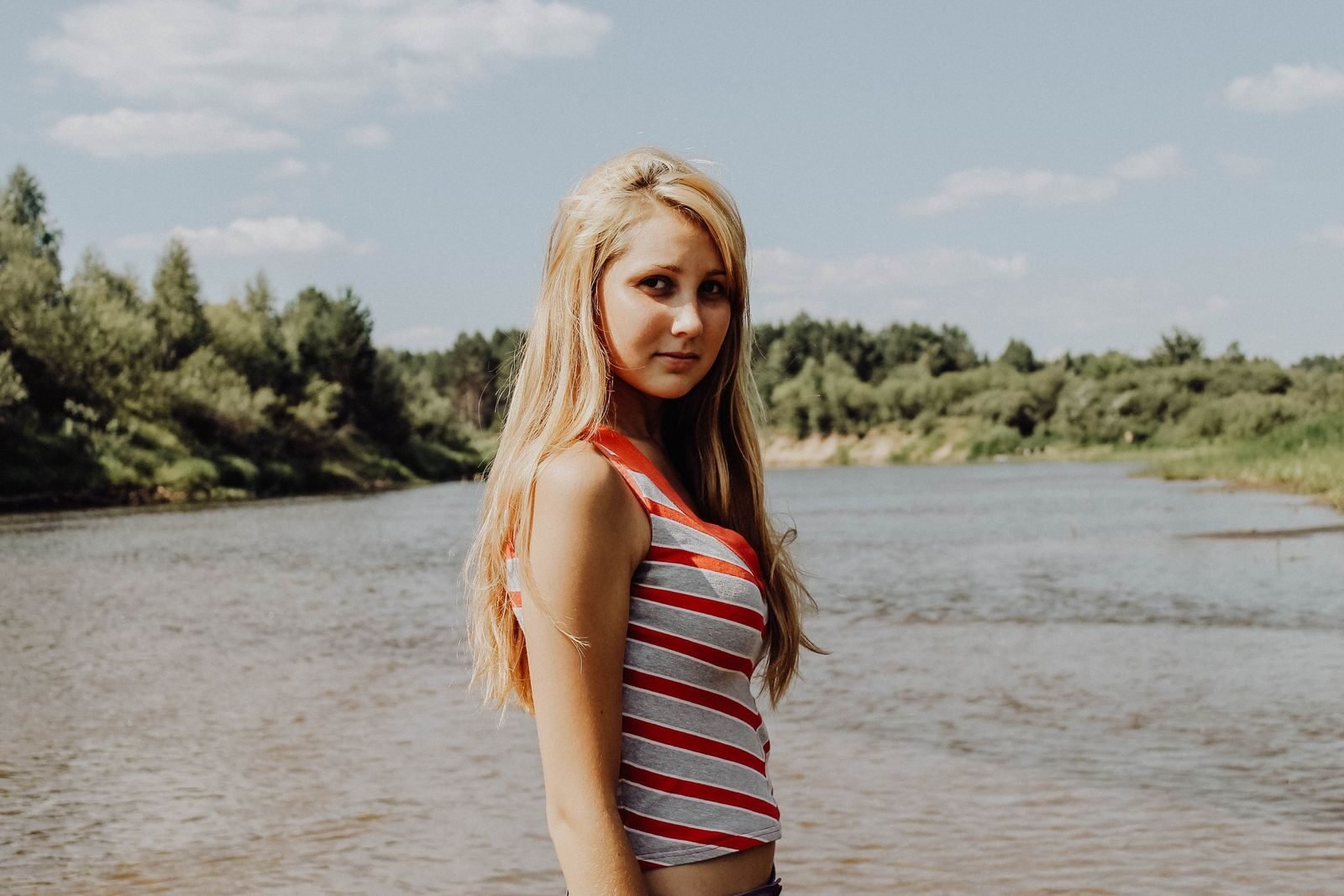 Blonde woman in striped top stands by a river on a sunny day outdoors.