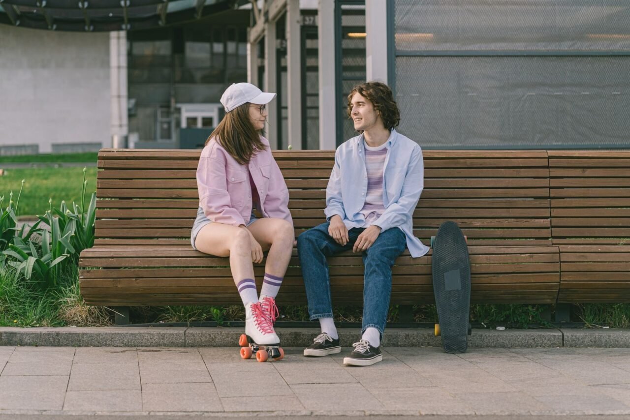 Caucasian man and woman sitting on a bench, talking with skateboards nearby outdoors during the day.