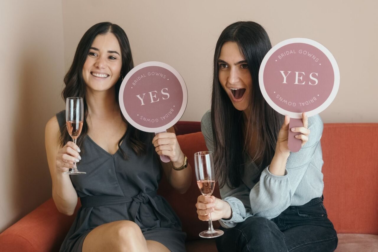 Two women celebrating a bridal fitting with champagne and 'Yes' signs.