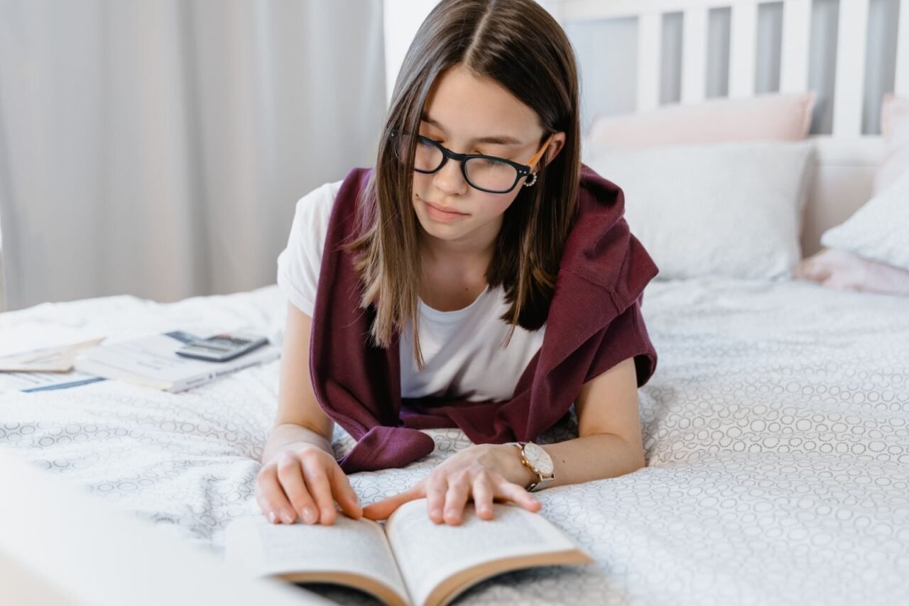 Teen girl with glasses reading a book on a cozy bed, emphasizing learning and relaxation.