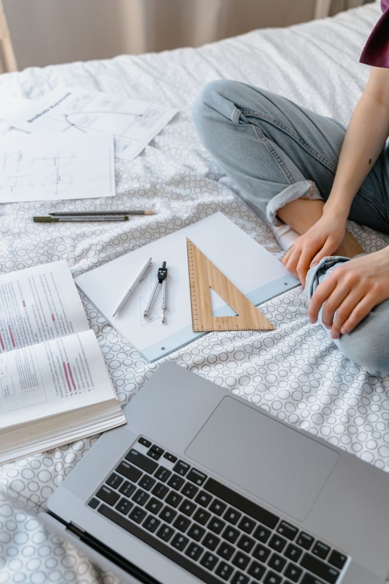 A student sits on a bed working with notebooks and a laptop, focusing on geometry homework.