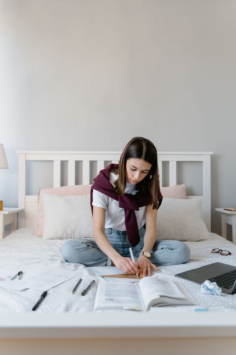 Teenage girl focused on studying at home, sitting on bed with open books and laptop.