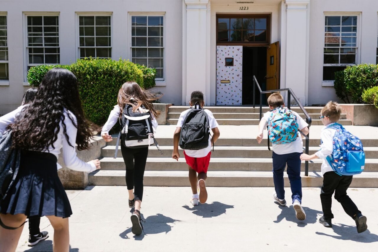 Group of students with backpacks running into school, captured from behind, showing excitement.