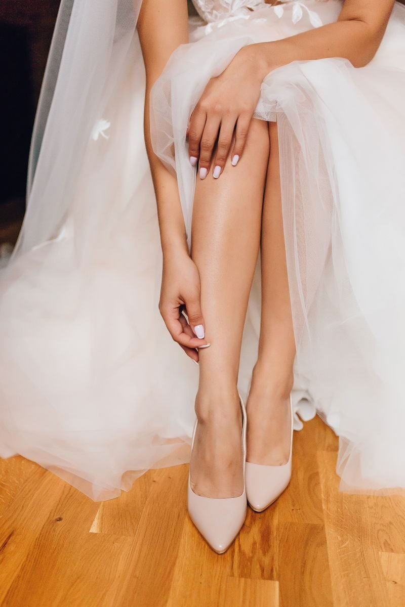 A bride in a tulle dress elegantly adjusts her high heels, capturing a moment of wedding preparation.