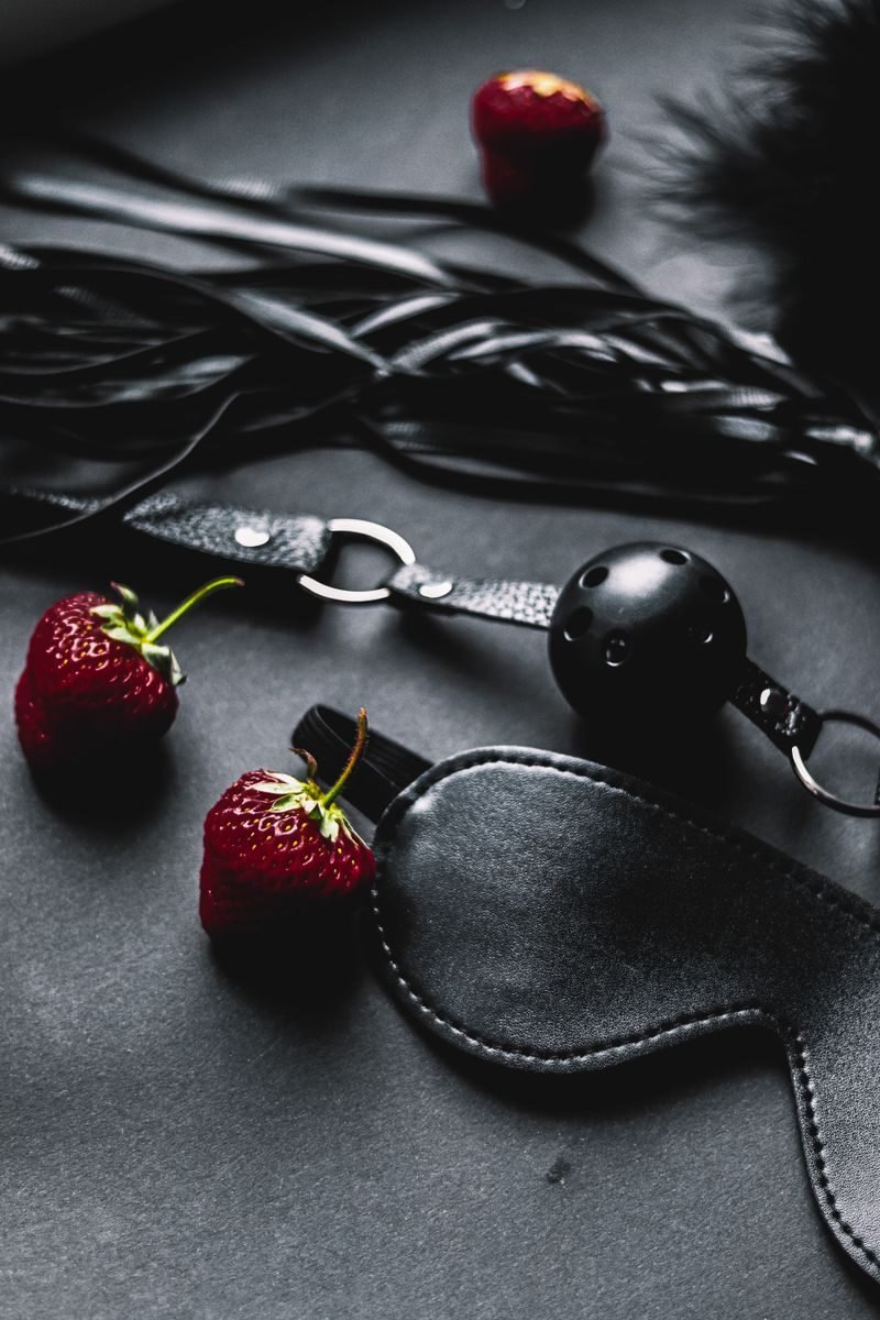 Close-up of erotic accessories including leather and strawberries against a black background.