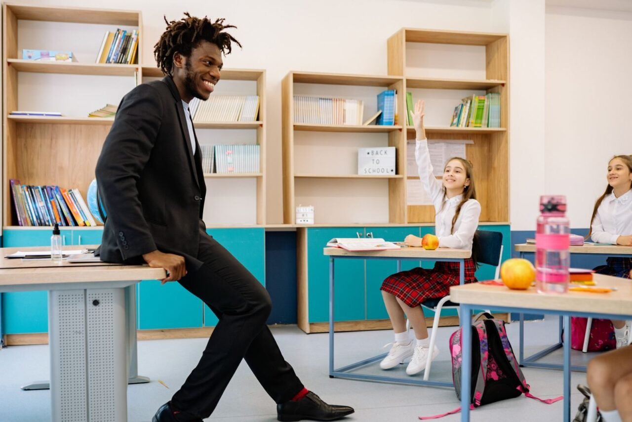 Teacher in classroom with students raising hands, engaging in lively lesson.