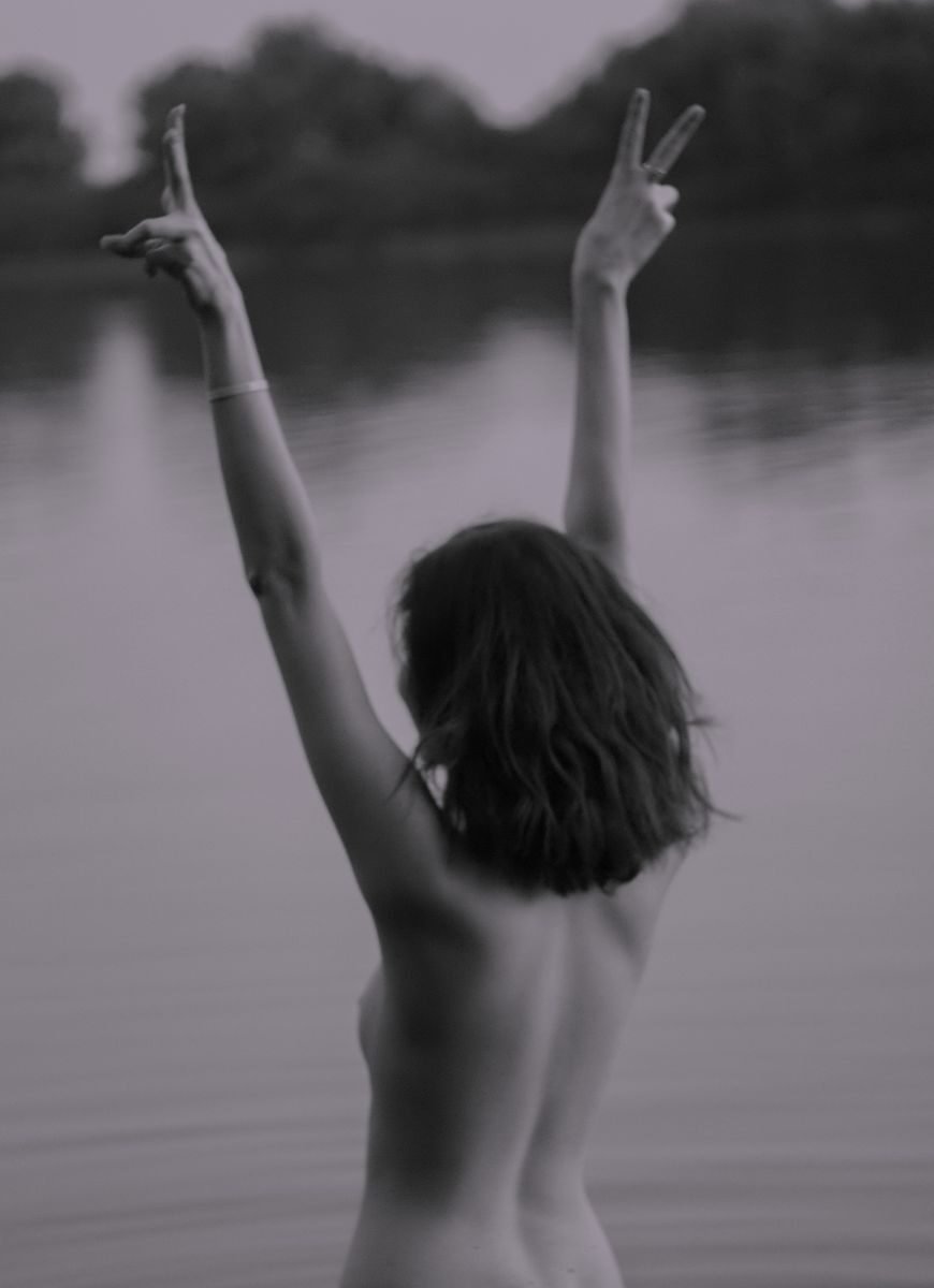 Monochrome image of a nude woman with arms raised, posing near a tranquil lake.