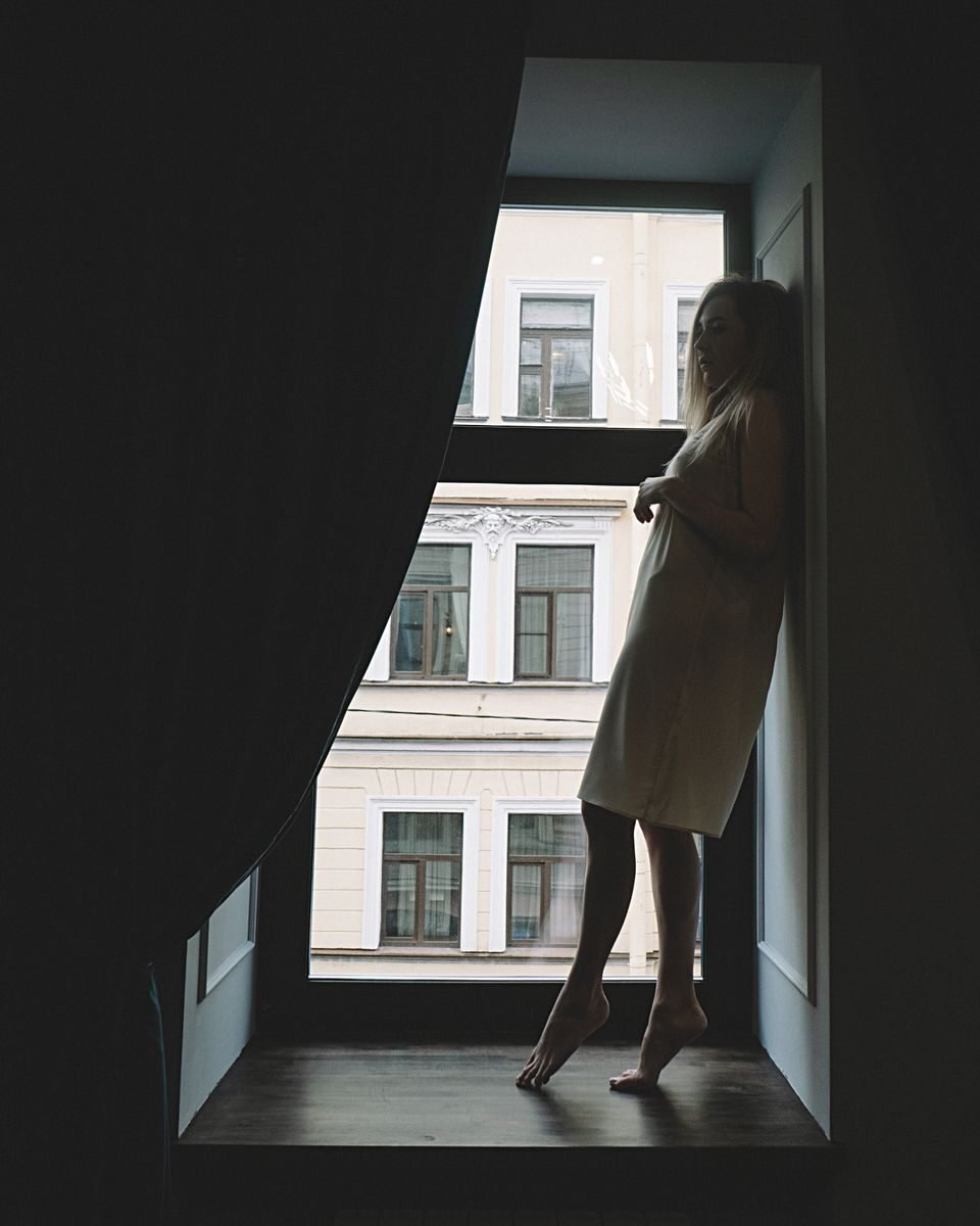 Silhouette of a young woman standing and leaning by a large window, with curtains framing the view.