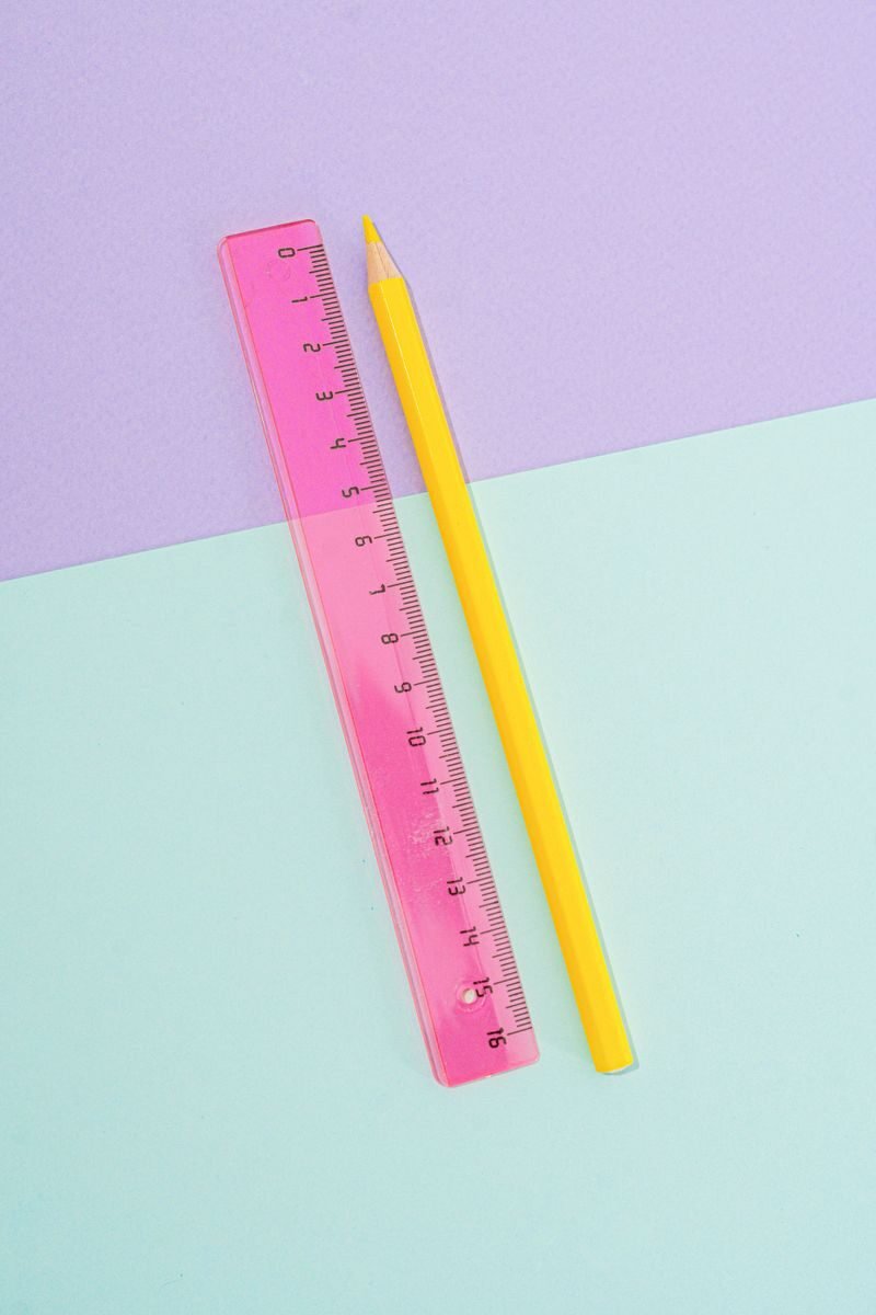 A bright yellow pencil and pink ruler on a pastel purple and green backdrop.