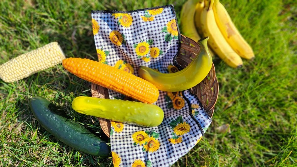 A basket on the ground with three silicone dildos in it, shaped like a cucumber, banana, and corn on the cob. In the grass surrounding the basket, there are actual fruits and veg.