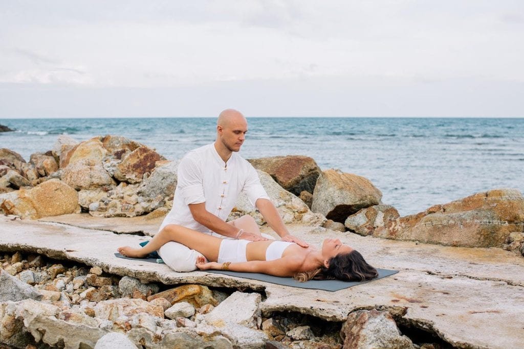 Moteris tantros poza guli ant jogos kilimėlio, rankas švelniai padėjusi ant krūtinės.