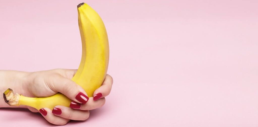 Display image for "how to give joi" article. A person with painted red fingernails grips a bright yellow banana in front of a pale pink background.