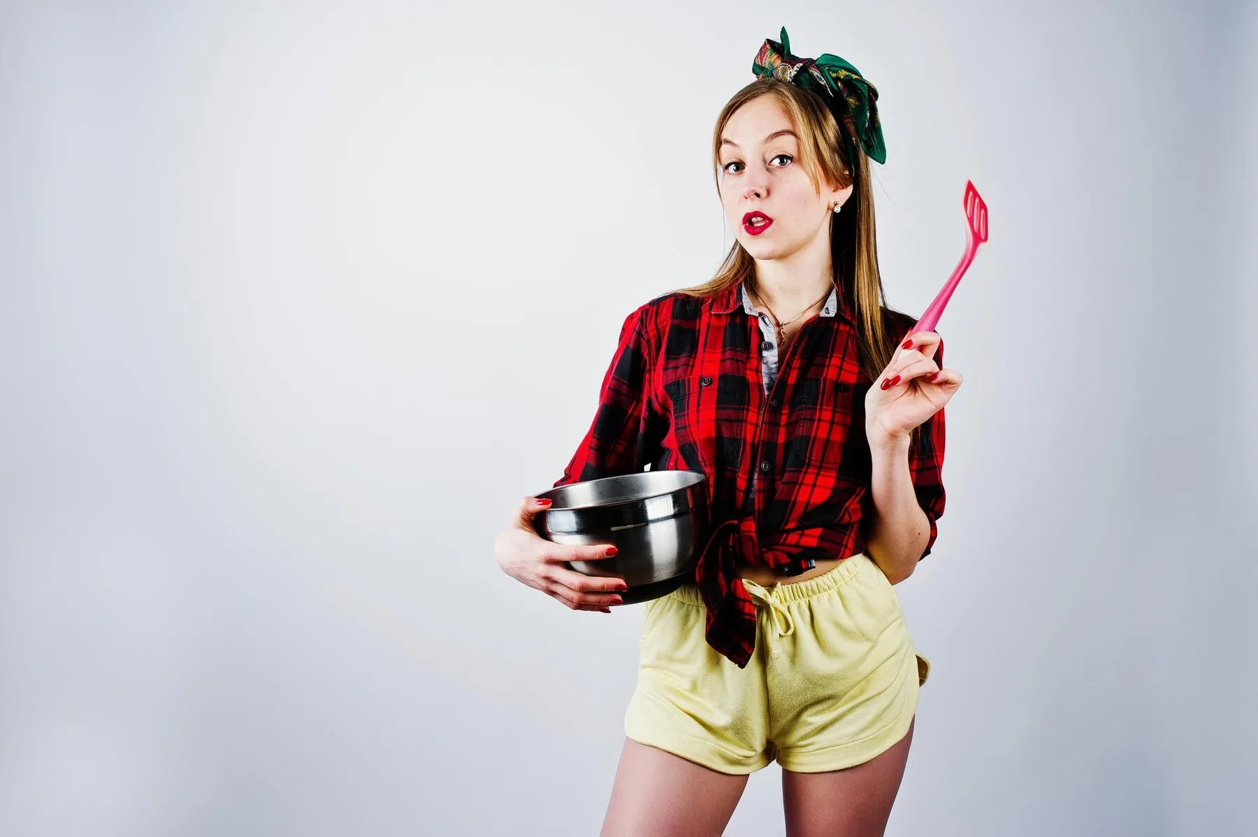 Young funny housewife checkered shirt yellow shorts pin up style with saucepan kitchen spoon isolated white background